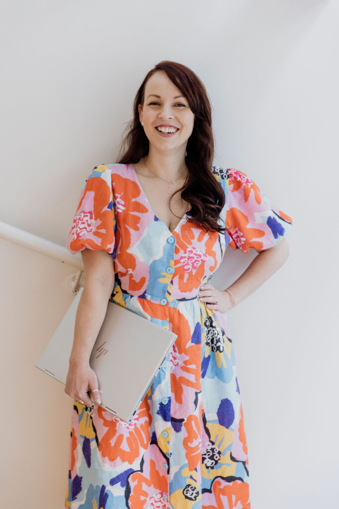 Dr Natalie Barre, productivity expert at Live an Intentional Life.  She is standing in a colourful dress holding her computer.