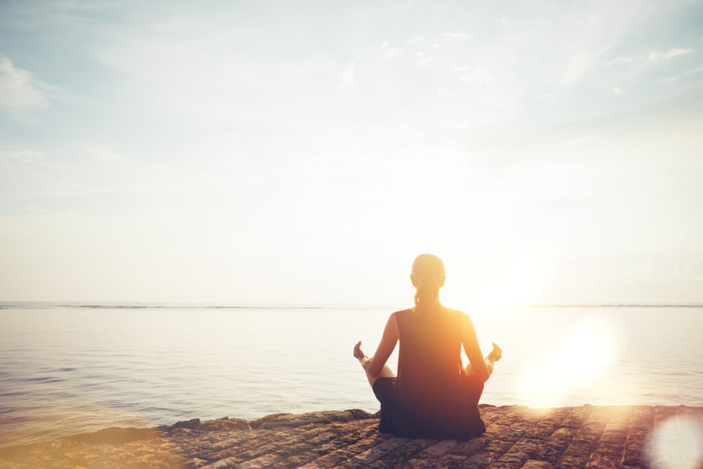 A woman meditating by the ocean in the sunlight