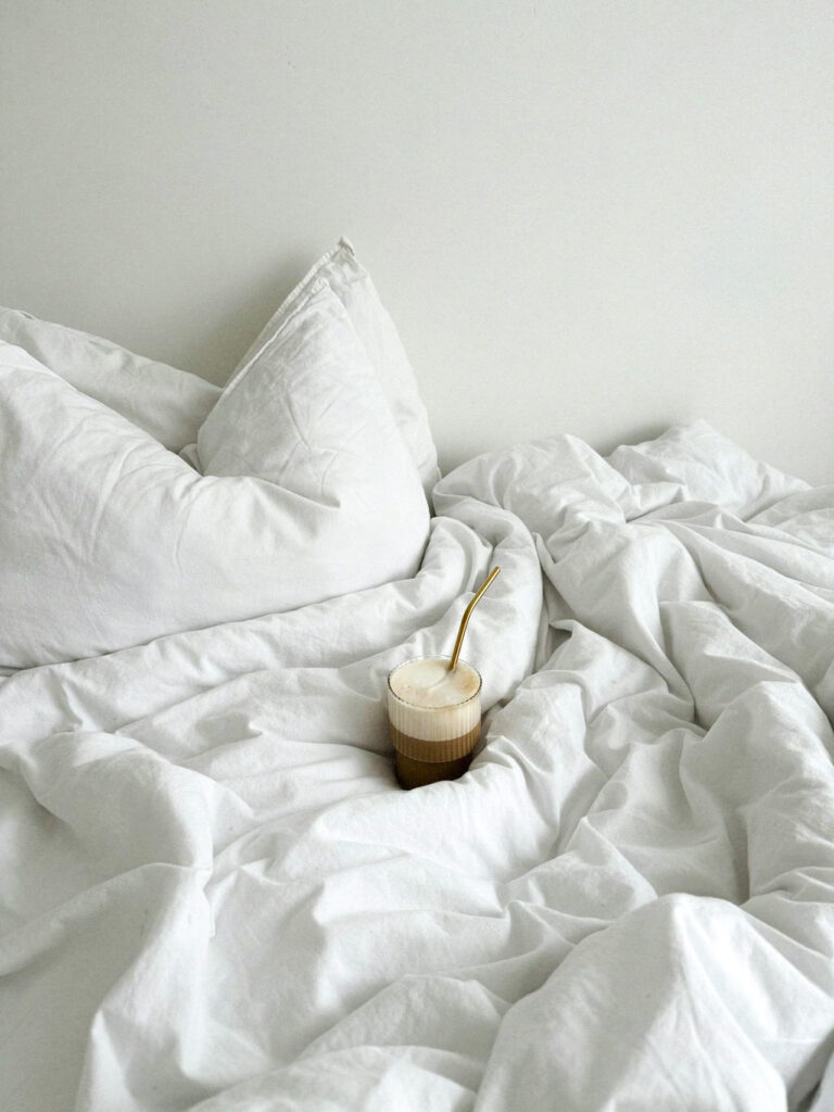 A creamy drink sitting on a bed with a white blanket and white pillow.