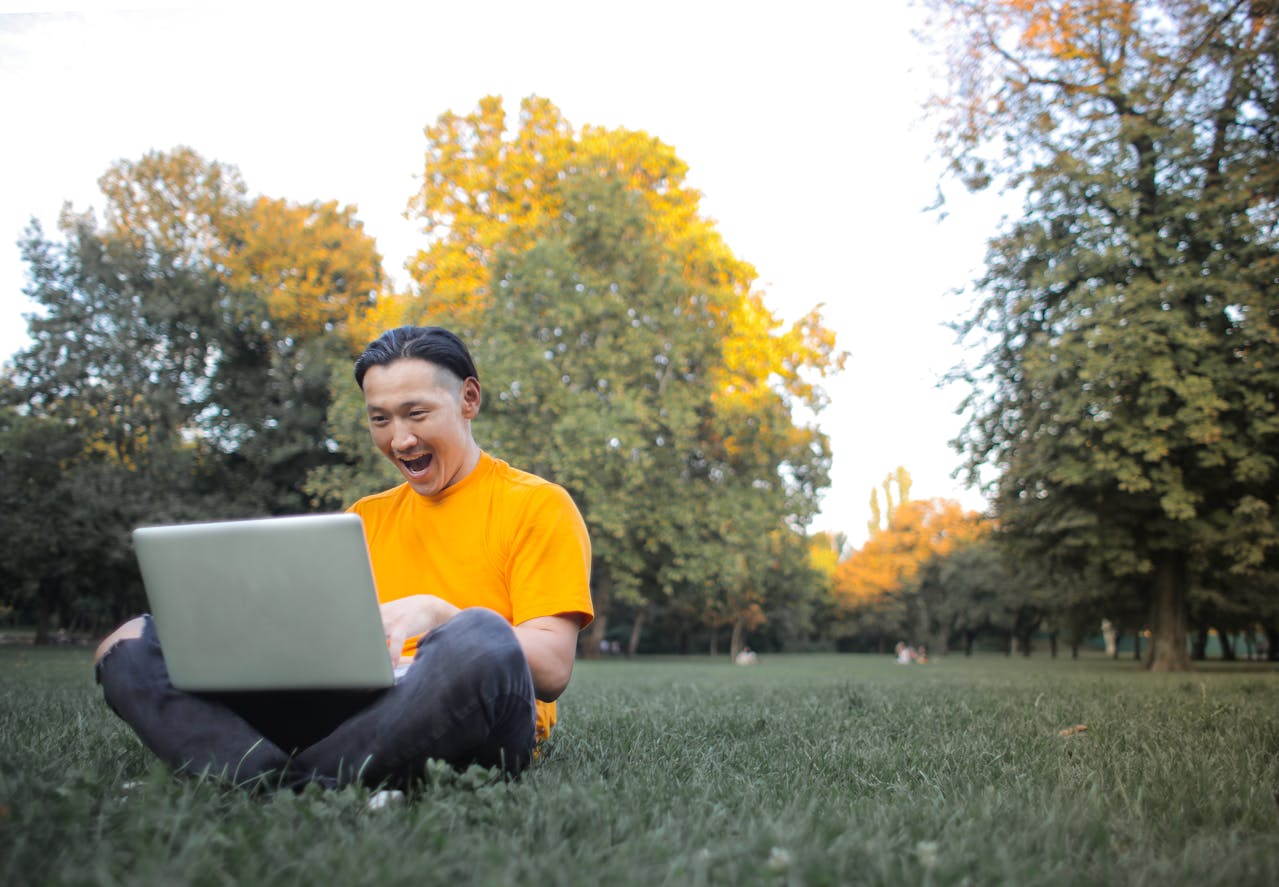 A man in a yellow tshirt sitting on the grass. He is looking at his laptop with an excited look on his face.
