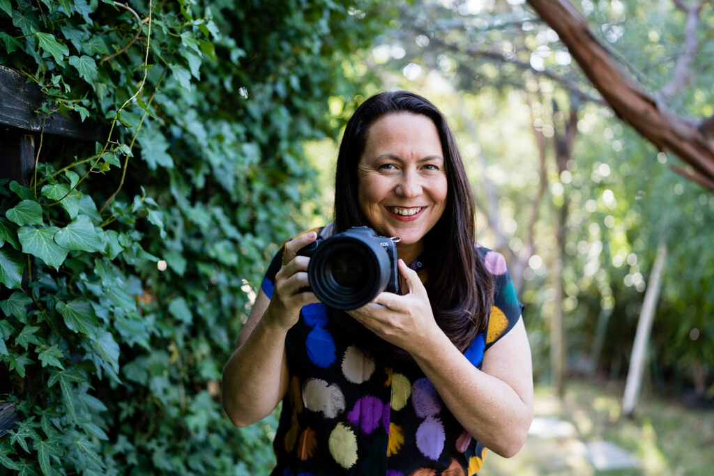Sharne Perrett is holding her professional camera and standing in front of a hedge.